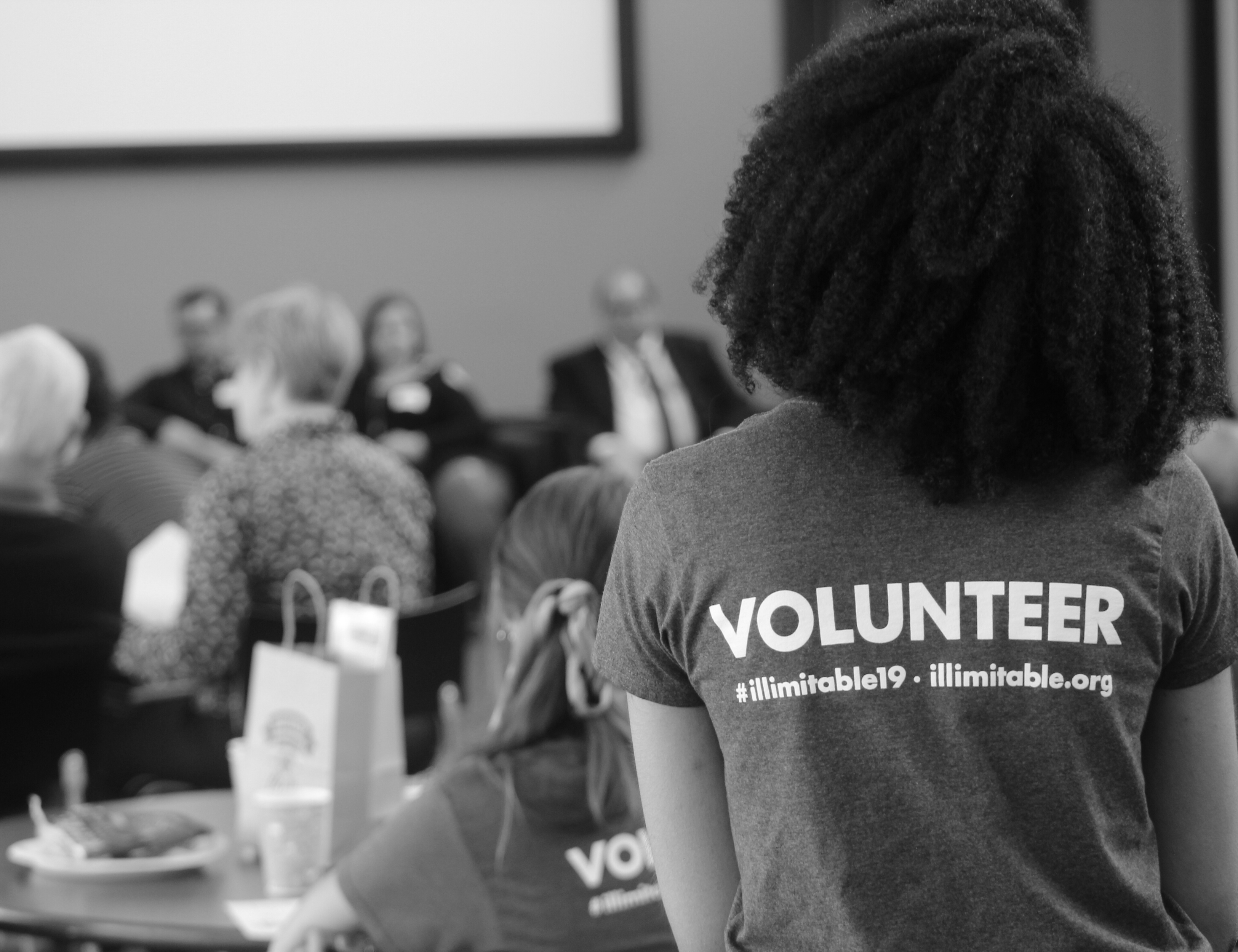 An African-American woman wearing an Illimitable tee shirt faces away from the camera. A group of panelists, the audience, and gift bags from vendors are visible in the background.